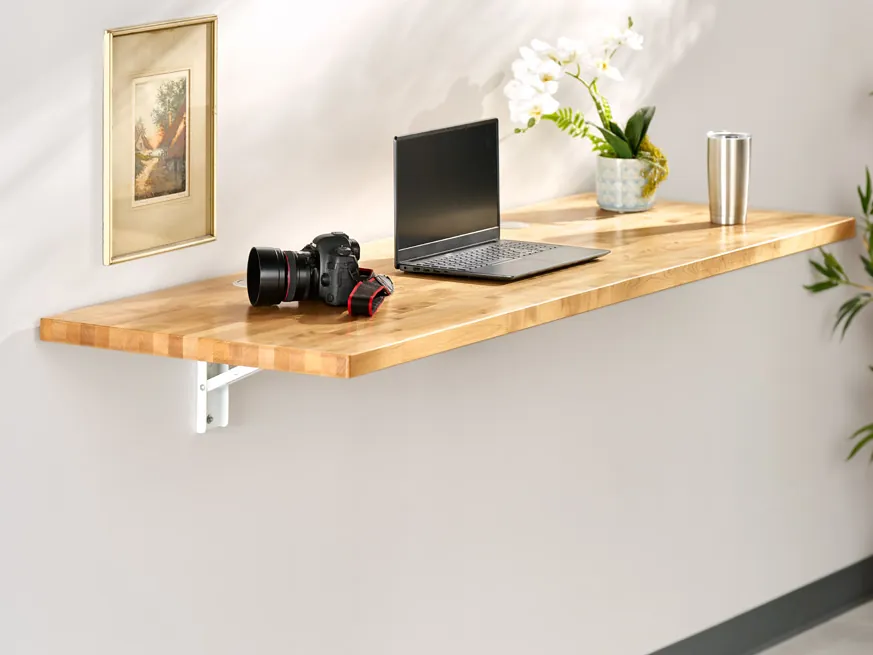 UPLIFT Floating Desk open on a wall, its work surface held level by white fold-down steel brackets, with a laptop and a camera on top (shown with a butcher-block top)